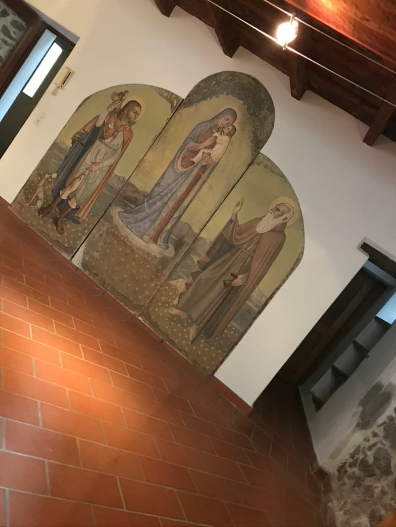 The image shows three wooden panels with paintings of religious figures mounted on a white wall above a red brick floor. There is an entrance in the background and a lamp on the wooden ceiling.