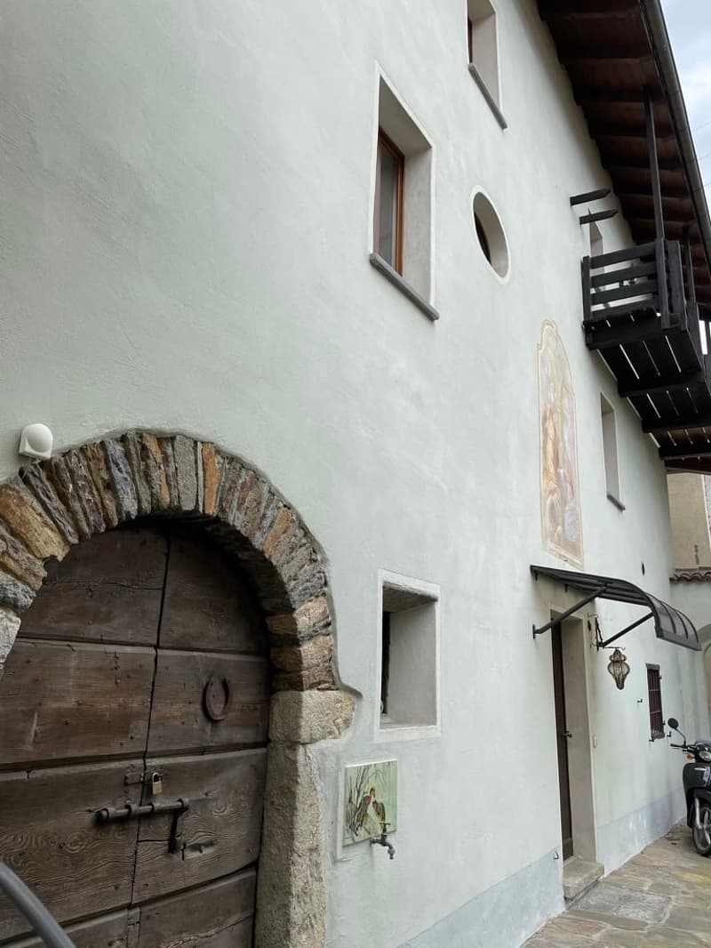 Facade of a white stucco building, wooden arched door, balcony with wooden railings, round window, mural on the wall