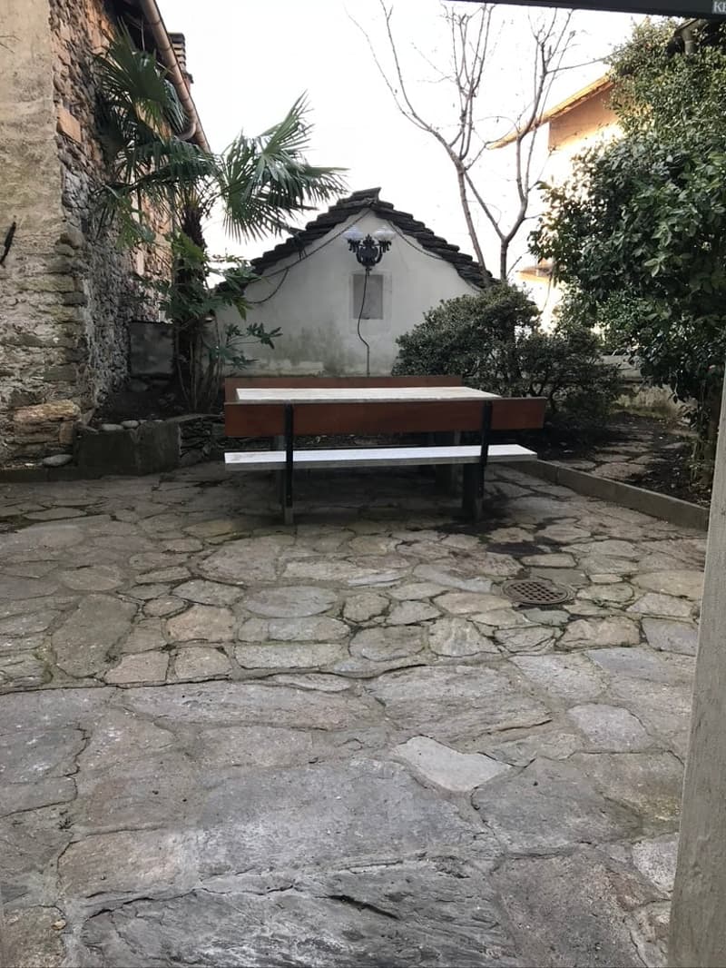 Garden with a table and benches, stone flooring, surrounded by plants and trees, small house in the background.