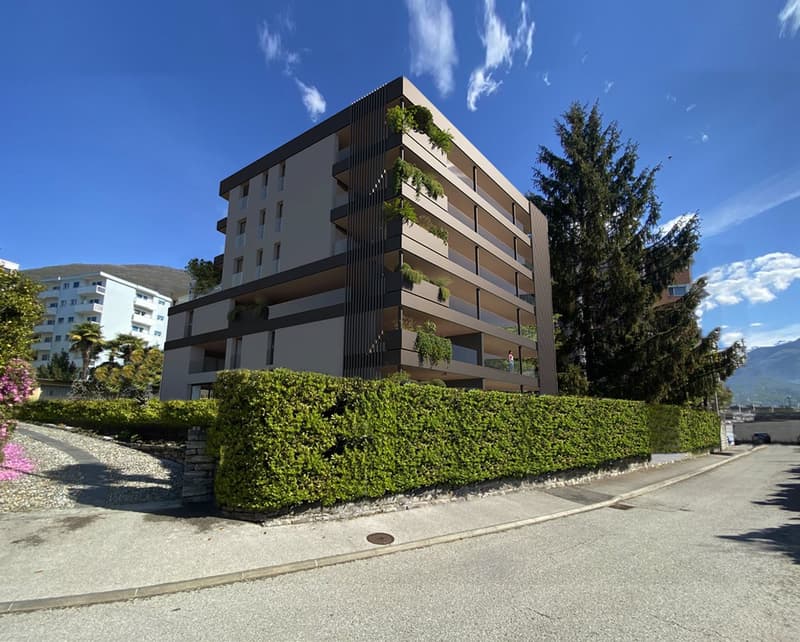 A modern multi-story residential building with balconies, surrounded by greenery and mountains