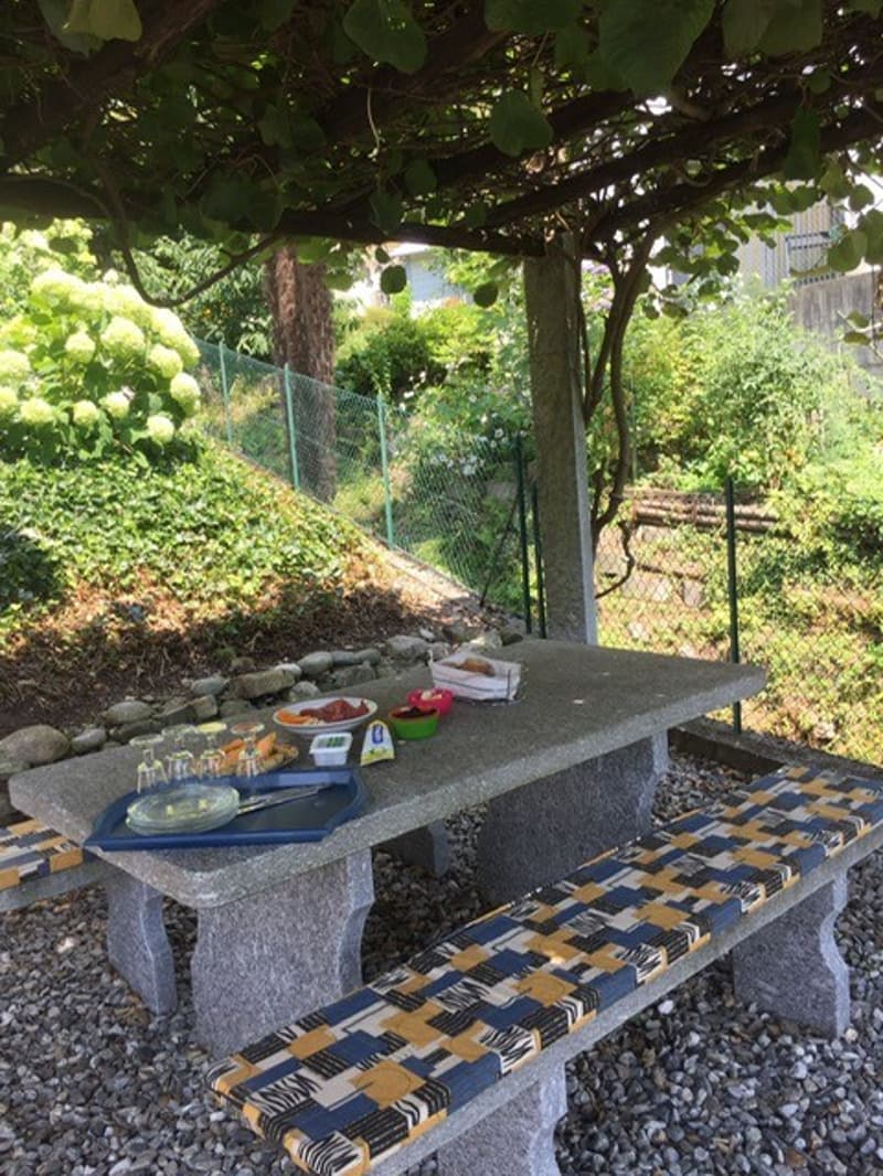 Outdoor dining table and bench, table with glasses and snacks, stone bench, gravel ground, greenery, and plants