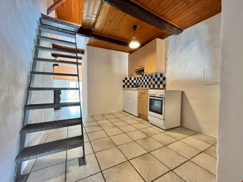 The picture shows an interior of a kitchen with white tiled floor, white walls, wooden exposed beams, white oven, white refrigerator, wooden cabinets, a staircase, and a light hanging from the ceiling.