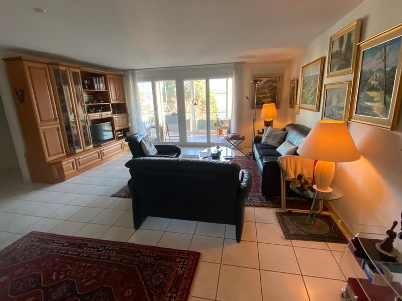 Living room with white tile flooring, wooden cabinets, glass walls leading to a balcony, black leather sofas, glass center table, carpet, lamps, framed pictures
