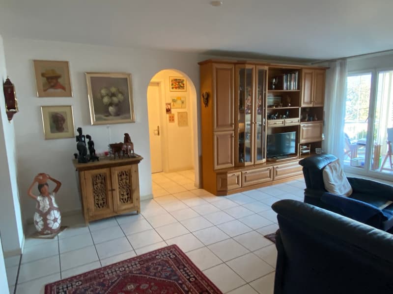 living room with white tiles, wooden TV cabinet, two armchairs, TV, decorative items, rug, picture frames, open door to another room