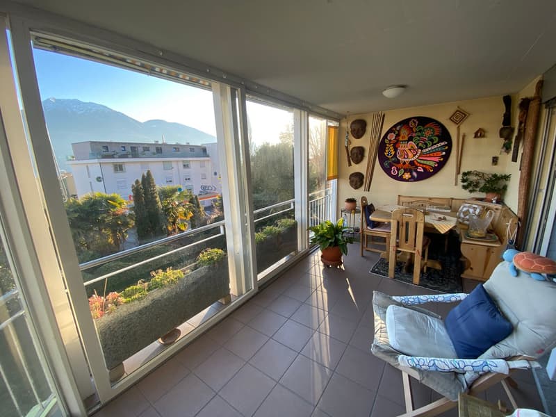 large balcony with panoramic mountain views, glass doors, wooden table with chairs, plants, cushion, lamp, wall decorations