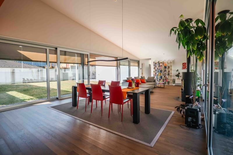 Dining room with large windows, wooden floor, a large desk with red chairs, a couch with cushions and a bookshelf in the background.