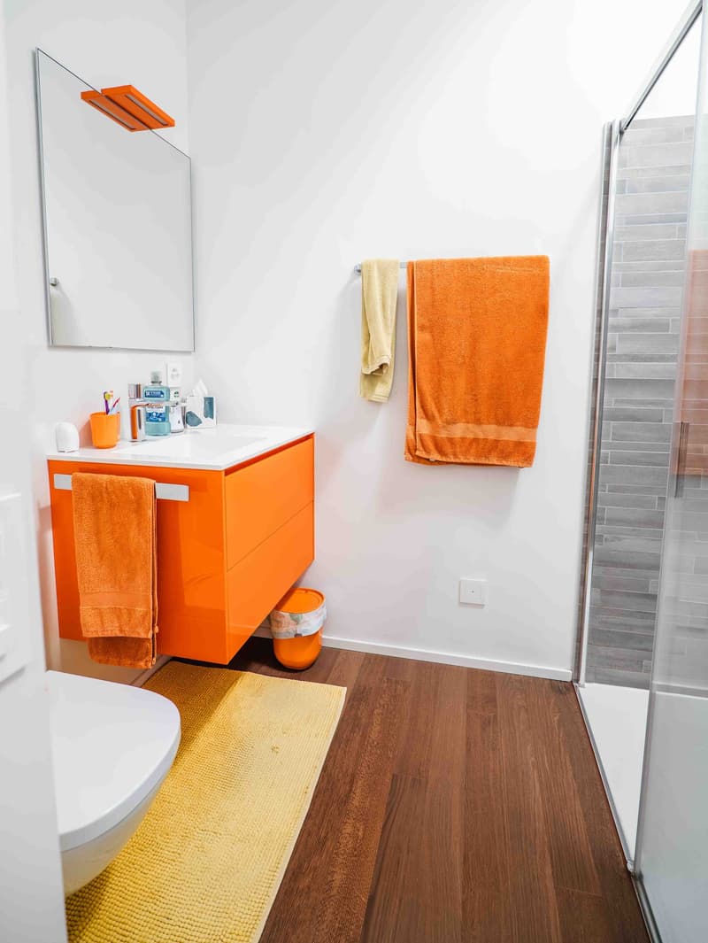 Orange cabinet with sink, white porcelain toilet, glass door shower enclosure, yellow rug, mirror, wall-mounted towel rack with towels