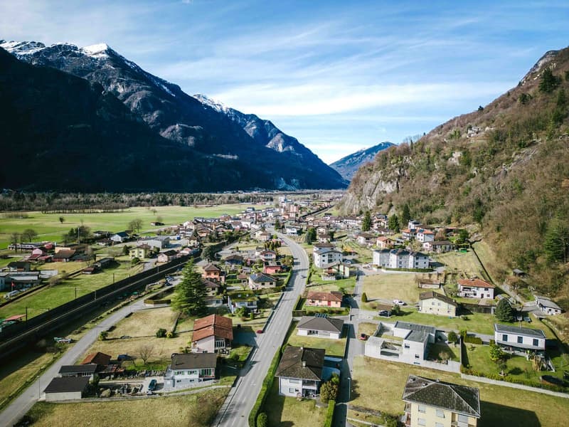 A village nestled in a valley surrounded by mountains, with a main road, several houses, and a vast green field.