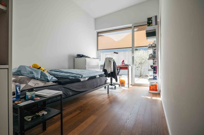 A room with a bed, bedside table, desk, chair, cabinets, books, shoes and bags on a shelf