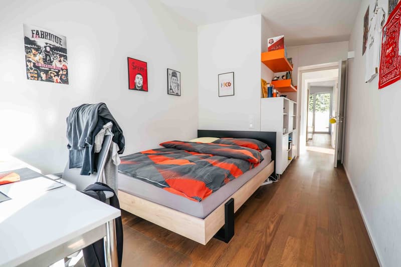 double bed with two pillows and duvet, wooden desk with a book on top, clothes hanging from chair, two posters on wall, hallway leading to white cabinet and hallway to other rooms