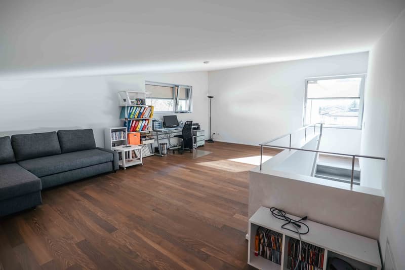 Wooden floor, white walls, gray couch, bookshelf, desk with monitor and chair, lamp, staircase with metal railing, window, white storage unit with books