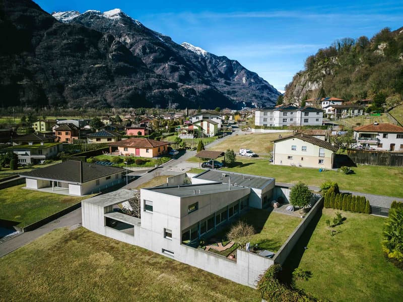 Large modern concrete house, nestled among houses, surrounded by mountains, valley, and greenery.