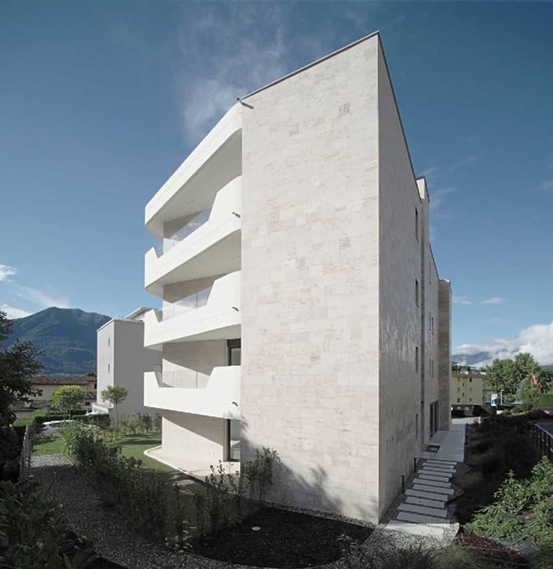 modern white stone building with balconies, three levels, steps leading to entrance, garden, trees, mountains in background