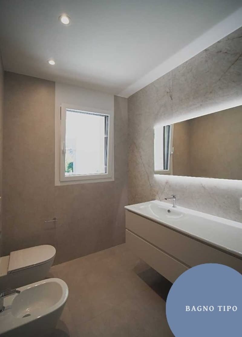 Spacious bathroom with white tile flooring and marble walls. Includes a double sink with mirrors, a modern shower enclosure, and a white toilet. There is also a small window providing natural light.