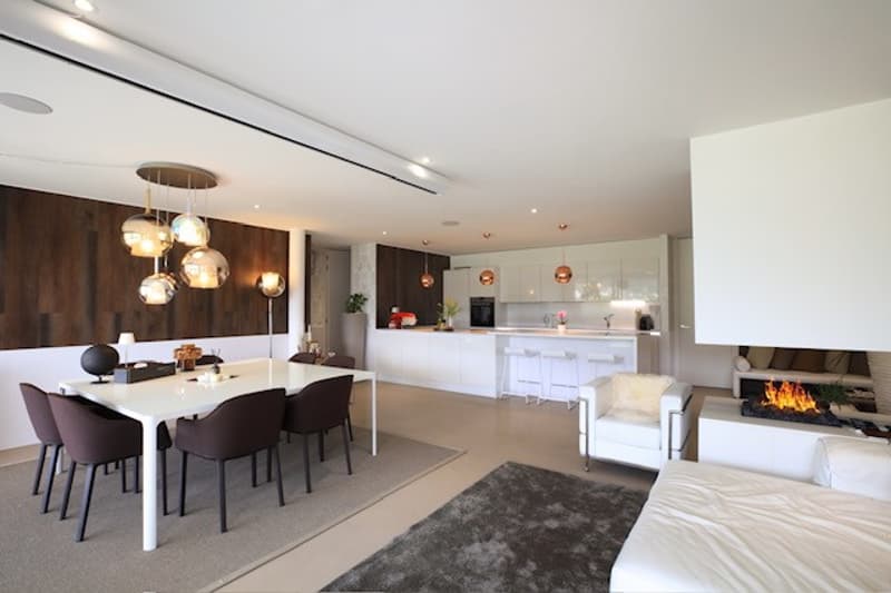 Fireplace, white table with chairs, beige carpet, white chair, large living room, white kitchen with modern cabinets, ceiling lamps, wooden wall
