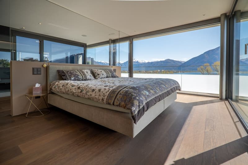 modern bedroom with large windows, wooden floor, bed with floral bedding, bedside table with tissue box, glass wall, mountain view