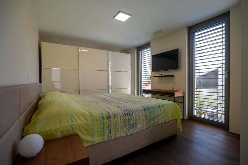 modern bedroom with large bed, large windows, wooden floor, white walls, ceiling lamp, bedside table, TV mounted on wall, sliding doors
