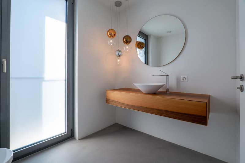 modern bathroom with white walls, wooden vanity, round mirror, white sink, faucet, glass door, and hanging lamps