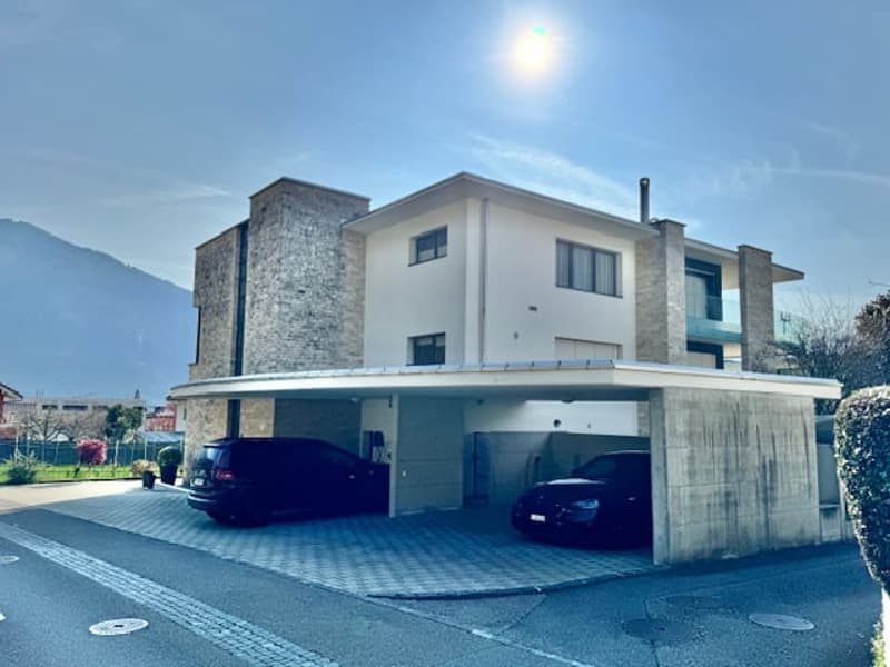 Modern two-story house with a garage, white exterior, two cars parked in the garage, stone wall on the left side, plants and trees in the background