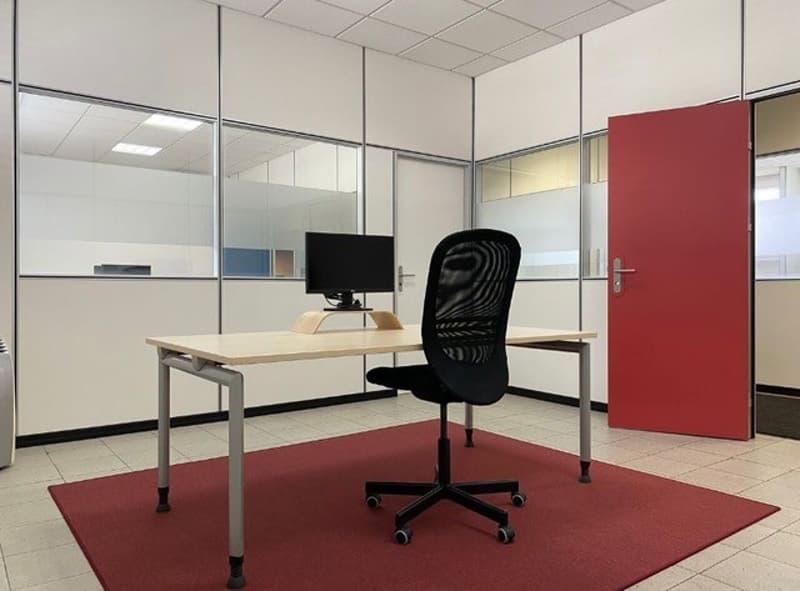 The image shows an office space with a desk, a computer monitor, and a black office chair. The walls are made of glass panels and there is a red door visible. The floor is tiled and there is a red carpet under the desk.