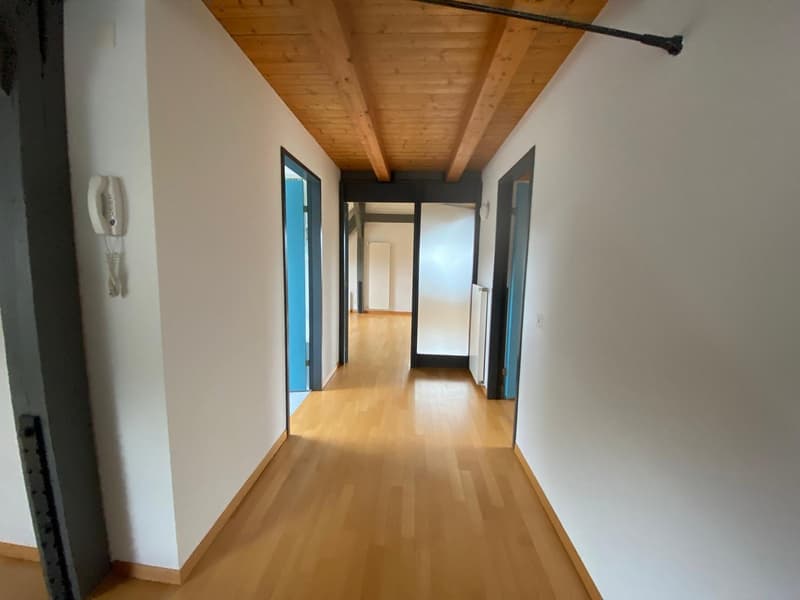The image shows a hallway with a wooden ceiling, white walls, and hardwood floors. The hallway has multiple doors leading to other rooms.