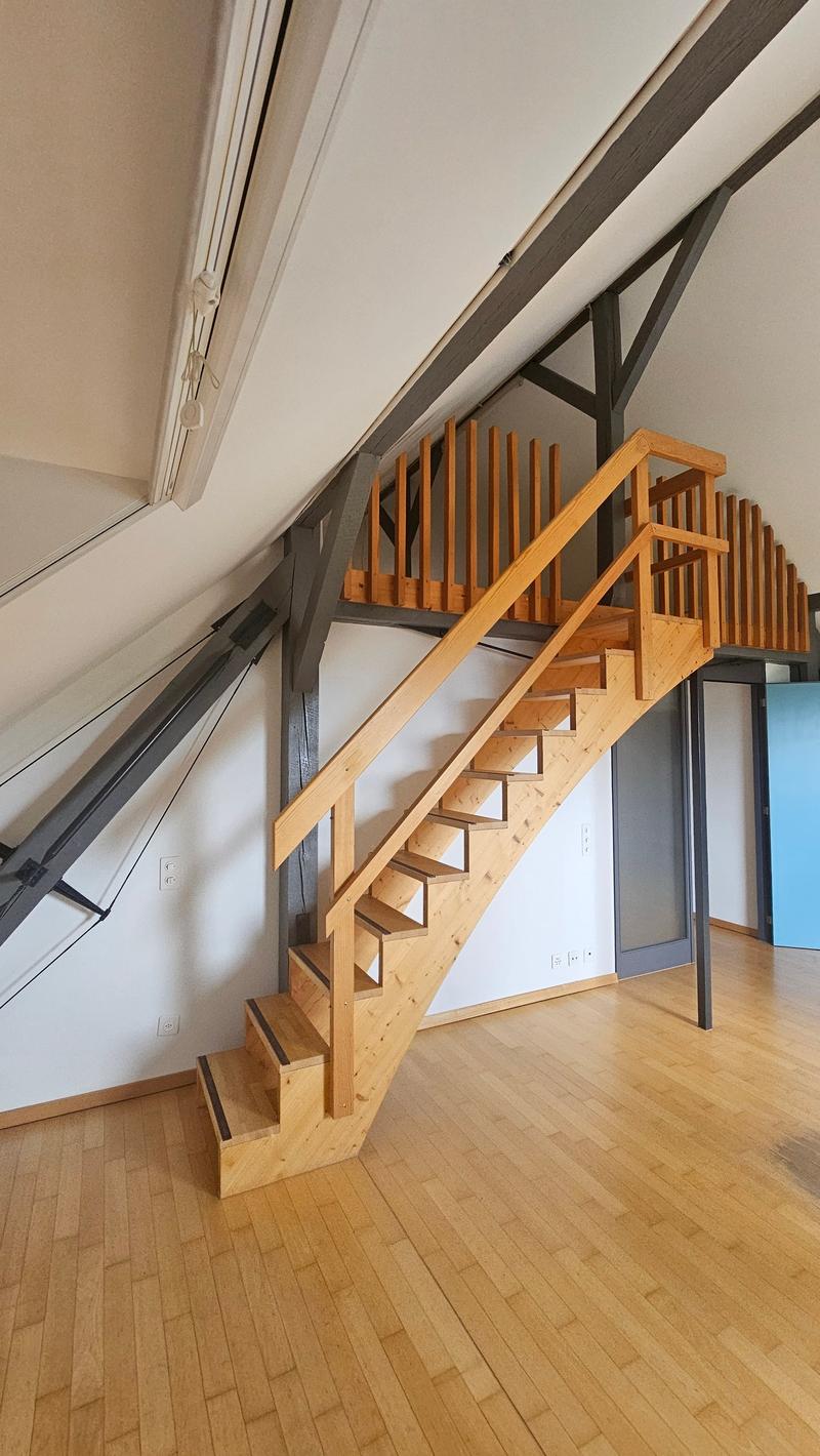 The image shows a wooden staircase with a modern, open design. The stairs have a natural wood finish and are supported by a metal frame. The staircase is located in a room with white walls and a hardwood floor.
