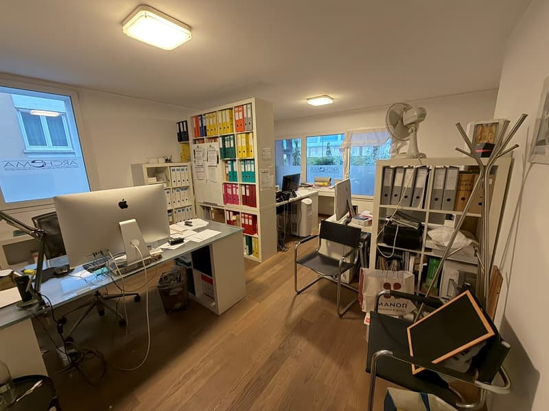 Office with wood floors, two Apple desktops, three chairs, desk with drawers, shelves, a fan and a file cabinet.