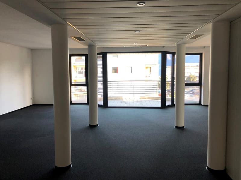 Empty room with white walls, black carpet, and four white pillars. Large windows with glass doors leading to a balcony.