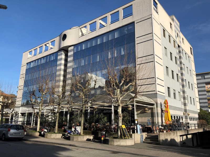 Modern building with glass facade, multiple windows, and a covered outdoor seating area with trees and umbrellas.