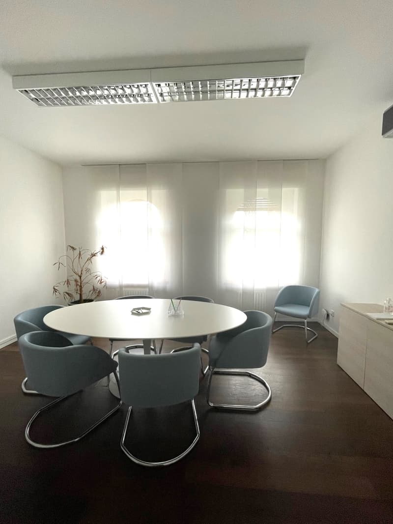 white round table, six chairs around, small bottle on table, white walls, white ceiling lights