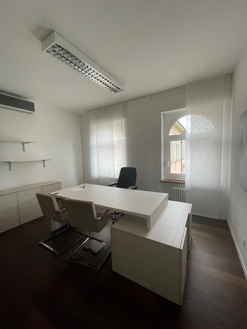 Office room with desk, chairs, shelves, and window with white curtain