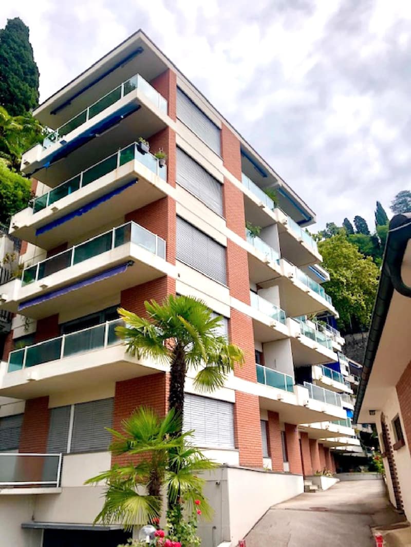 Modern multi-story apartment building with brick walls and glass balconies, surrounded by greenery, with a driveway in front