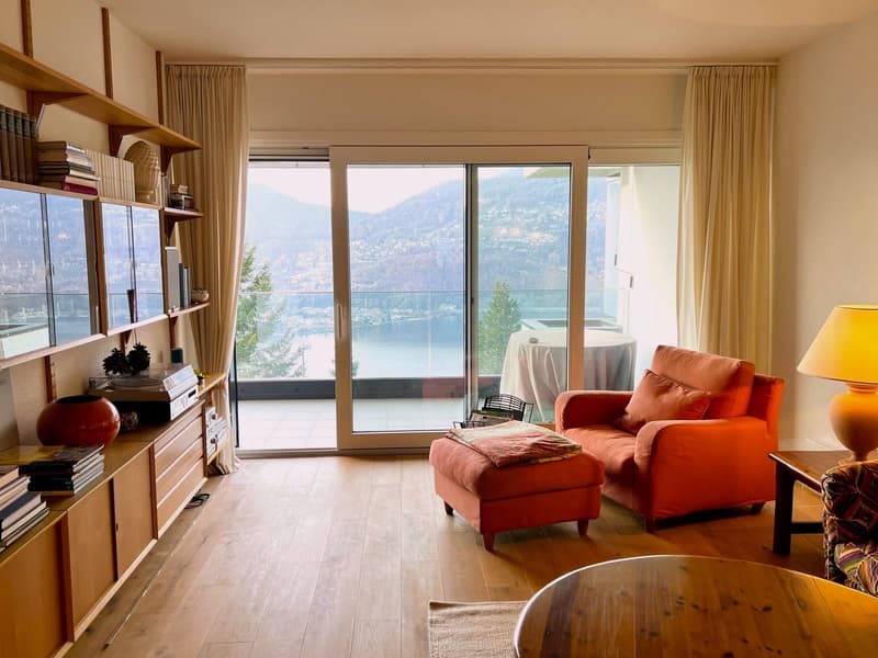 Living room with wooden floor, large windows, balcony, orange chair, ottoman, table, lamp, shelves with books, TV, and a view of the lake and mountains