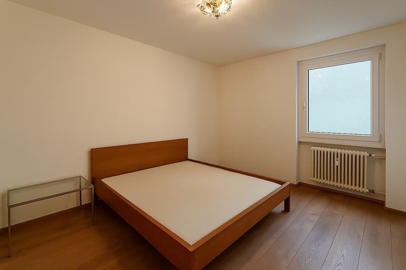 Empty room with wooden floor, white walls, bed, glass table, window, radiator
