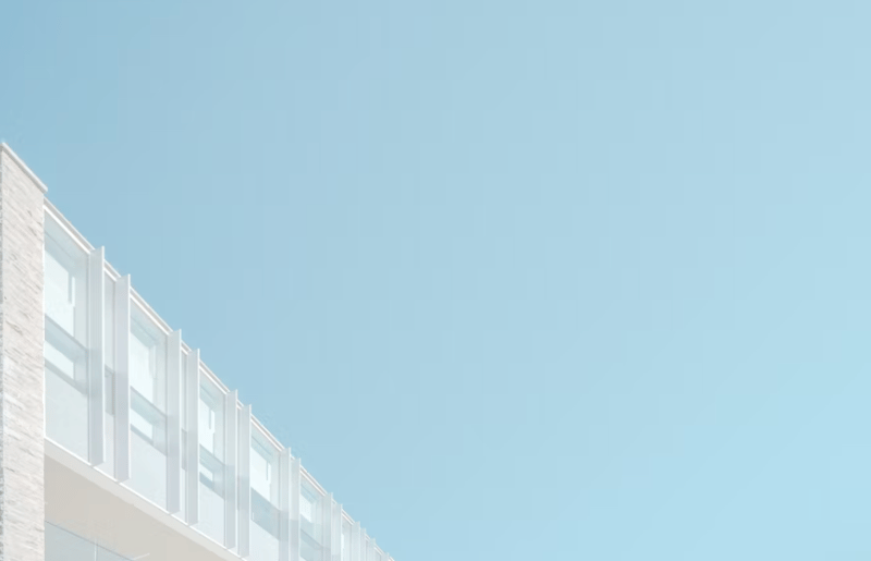 The image shows a modern, minimalist building with a series of large, rectangular windows arranged in a grid-like pattern. The building has a clean, white exterior and is set against a clear blue sky, creating a simple and elegant visual composition.