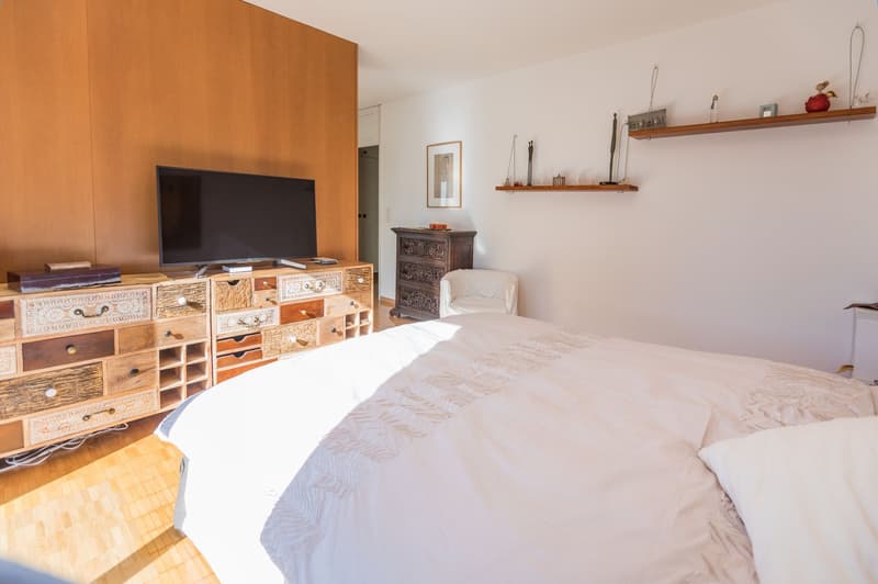 Large bed with white sheets and pillows, wooden chest of drawers with carved details, TV on top, small white armchair, picture frame on the wall, white walls, wooden floor.