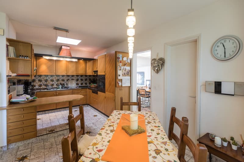 white walls, white floor, large wooden dining table, chairs, open cabinet with microwave, sink, wooden cupboards, wooden wall decorations, clock