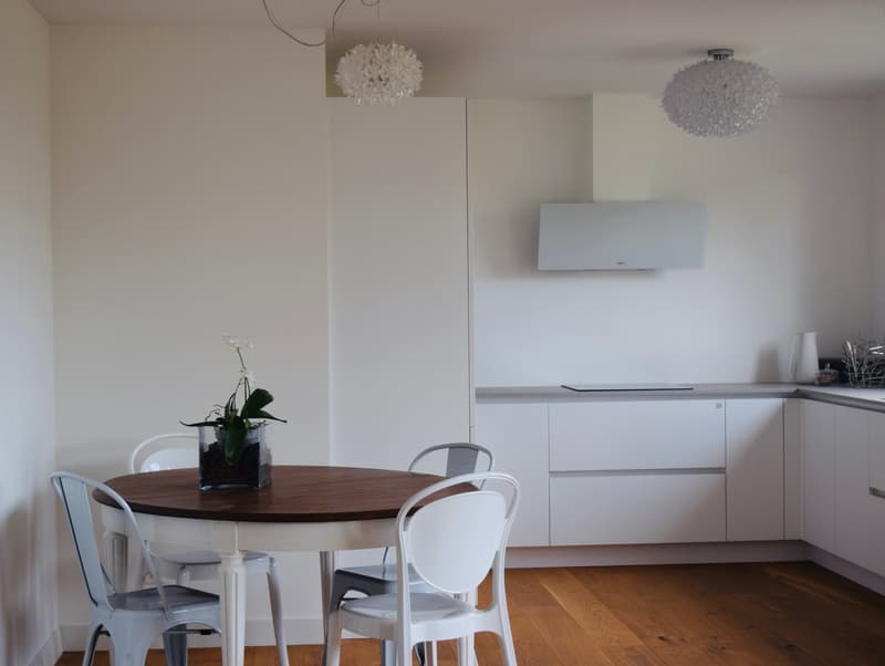Modern round dining table, white chairs, white walls, wooden floor, white cabinets, round lamps, kitchen area with stove