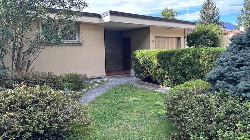 Single-story house with stucco exterior, stone accents, and a garage. The house is surrounded by lush landscaping, including trees, shrubs, and a well-maintained lawn. The entrance is covered by a small overhang.