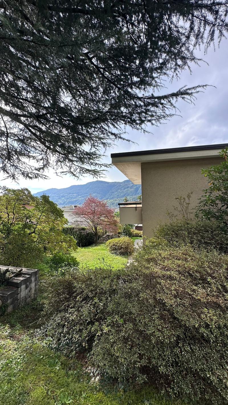 Two-story house with a modern design, surrounded by lush greenery and trees. The house has a balcony or terrace visible, and the landscape includes a garden with flowering plants and shrubs.