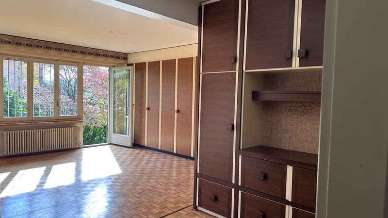Spacious living room with large windows overlooking a balcony, wooden built-in cabinets and shelves, hardwood flooring, and a cozy atmosphere.
