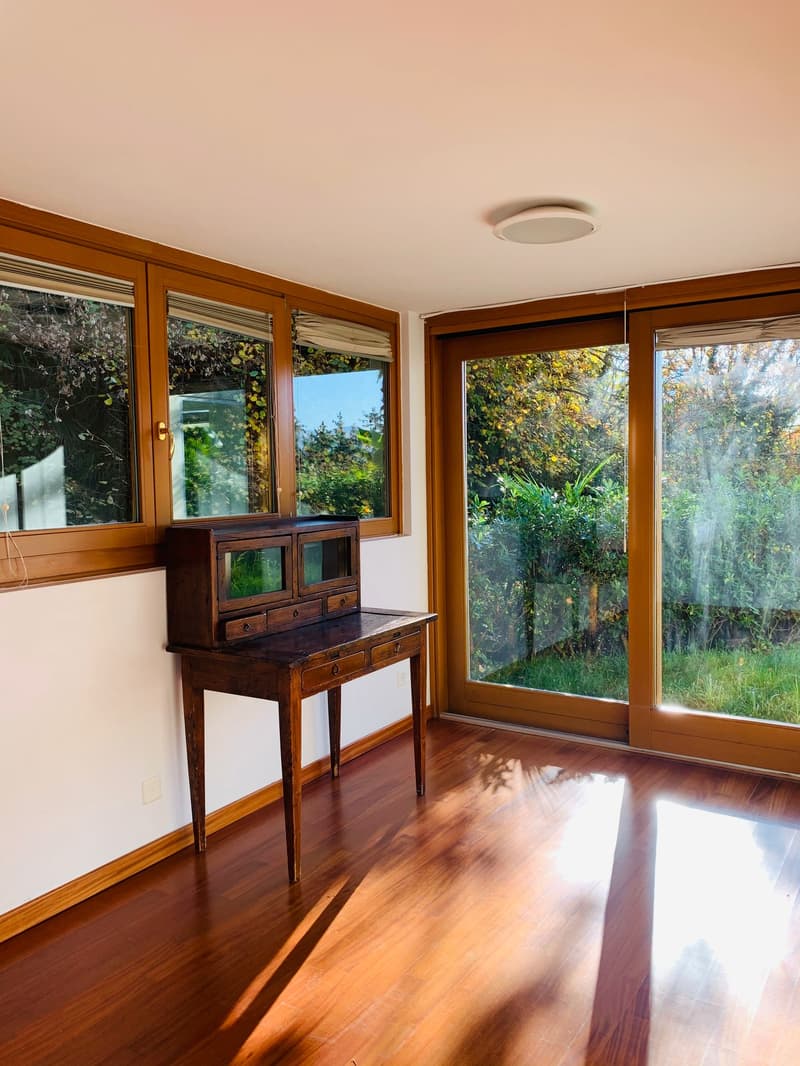 Room with wooden desk and wooden glass doors leading to a garden, large windows, wooden floor.