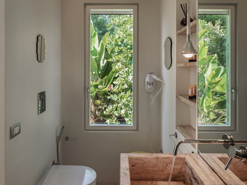 modern bathroom with white toilet, large window with view of greenery, double sink, shower head, shelves, and a lamp
