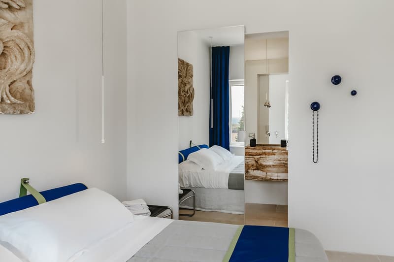 modern bedroom with white walls, blue and white bedding, mirror, artwork, window with blue curtains, wooden counter, bathroom