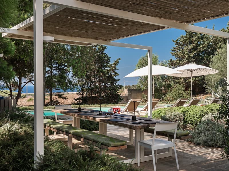 Outdoor dining area with wooden table and benches, covered by pergola, swimming pool, lounge chairs, umbrellas, ocean view