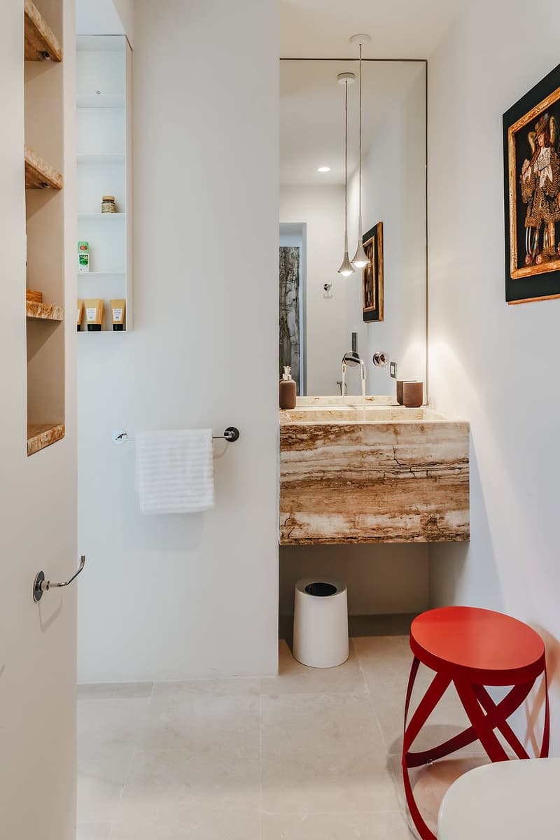 Bathroom with white walls, wooden shelves, a large mirror, a marble countertop sink, a red stool, and a white toilet.