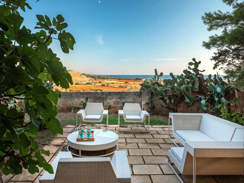 Outdoor seating area with a table, chairs, and a couch, surrounded by plants and a stone wall, with a view of the ocean and landscape.