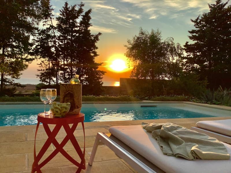 Outdoor area with a swimming pool, lounge chairs, a table with wine glasses, and a bottle of wine, surrounded by trees and a sunset view.