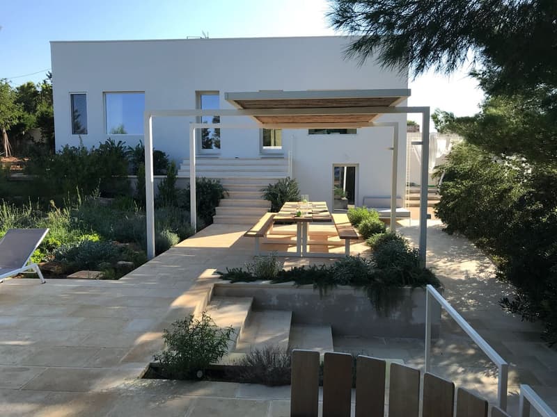 Outdoor dining area with wooden table and benches under a pergola, stone patio, white house, garden with plants and trees