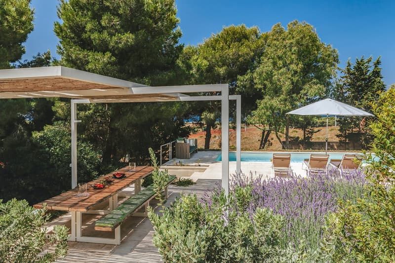 Outdoor dining area with wooden table and benches, pergola, swimming pool, lounge chairs, umbrella, lavender plants, trees